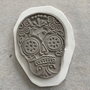 May include: A gray ceramic sugar skull with intricate floral designs and detailed eye sockets. The skull is on a white ceramic base.