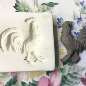 May include: A white silicone mold of a rooster, with a detailed design. The mold is next to a gray ceramic rooster, which is a finished product made from the mold.