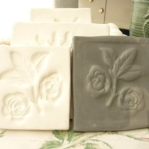 May include: Two square ceramic tiles with raised floral designs. One tile is white with a raised design of two roses and leaves. The other tile is gray with a raised design of three roses and leaves.