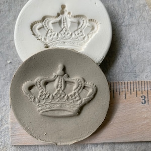 May include: A close-up of a white ceramic mold and a gray ceramic imprint of a detailed crown design. The mold and imprint are on a wooden surface with a ruler.