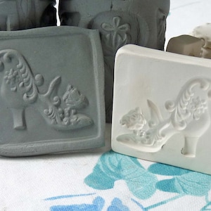 May include: Two ceramic molds for making decorative items. The molds are square and feature a raised design of a shoe with intricate details. One mold is gray and the other is white.