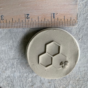 May include: A round, light brown clay coaster with a raised design of three honeycombs and a bee.