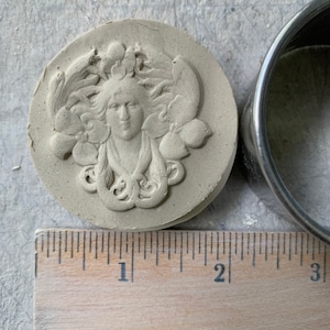 May include: A white ceramic mold with a raised design of a woman's face surrounded by leaves and fruit. The mold is approximately 2 inches in diameter.