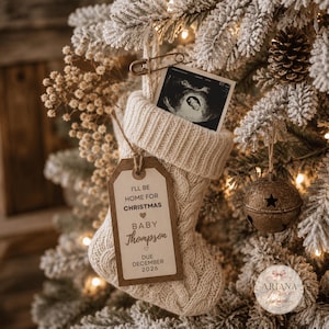 Peut inclure: Une chaussette de Noël en tricot torsadé couleur crème avec une étiquette en papier brun portant le texte I'LL BE HOME FOR CHRISTMAS BABY Thompson DUE DECEMBER 2026. Une échographie en noir et blanc est fixée en haut par une épingle à nourrice dorée.