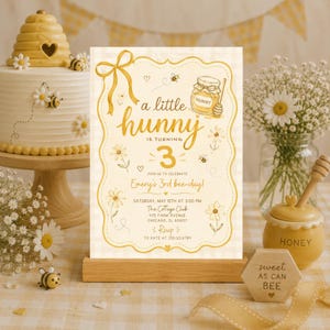 May include: A rectangular invitation card with a yellow gingham background, featuring a gold ribbon, a honey pot, and daisies. The text reads: a little hunny is turning 3, join us to celebrate Emery's 3rd bee-day, Saturday, May 15th at 3:00 PM, The Cottage Club, 475 Farm Avenue, Chicago, IL 60607, RSVP to Kate at 235.123.6789.