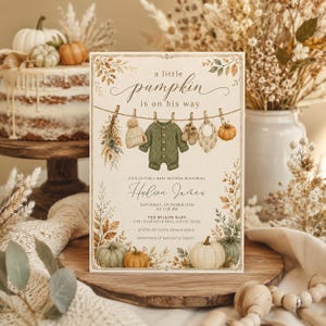 May include: A rectangular baby shower invitation card featuring the text 'a little pumpkin is on his way' above an illustration of baby clothes hanging on a clothesline, including a beanie, a green romper, booties, and a bib, surrounded by pumpkins.