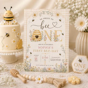 May include: A charming bee-themed first birthday party invitation with a matching cake and cookies. The invitation features the text "So Sweet to Bee One" and details for the event. The cake is decorated with bees and daisies. A honey dipper and decorative beads are also present.