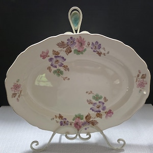 May include: An oval, off-white serving platter with a scalloped edge, adorned with floral patterns in pink, purple, and green. The platter is displayed on a white metal stand with a decorative handle. The floral design includes brown leaves.