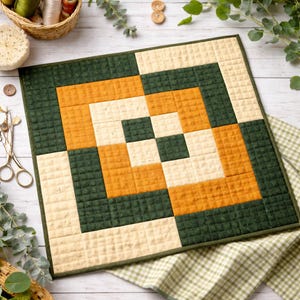 Op de afbeelding: Een vierkante, gewatteerde placemat met een geometrisch ontwerp. Het ontwerp bevat vierkanten in crème, mosterdgeel en donkergroen, gerangschikt in een symmetrisch patroon. De placemat is afgezet met een donkergroene stof en heeft een getextureerde uitstraling.