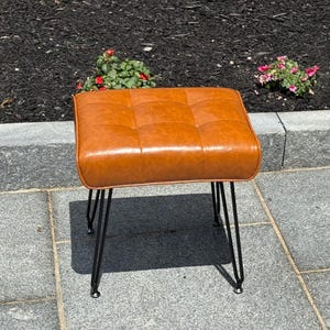 May include: A square, tufted ottoman with a warm brown leather-like top. The ottoman has a grid pattern and is supported by four black hairpin legs. The piece is outdoors on a stone patio.