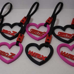 May include: Black and pink heart-shaped keyrings with red car model names: GTI, 350Z, STI, and WRX. Each keyring has a black strap and red connector.