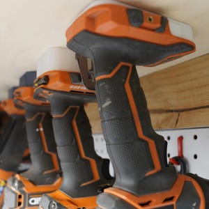 Könnte beinhalten: Eine Reihe orange-schwarzer Akku-Bohrmaschinen mit weißen LED-Leuchten, die unter einem Holzregal montiert sind. Die Bohrmaschinen sind in einer Reihe angeordnet, ihre Griffe zeigen nach vorne. Eine Lochwand und eine Schleifscheibe sind im Hintergrund zu sehen.