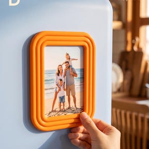 Peut inclure: Un cadre photo magnétique orange rectangulaire avec un design nervuré et superposé. Le cadre contient une photographie d'une famille sur une plage. Le cadre est fixé sur une surface bleu clair. Le texte indique : B, You imagine it, We print it.