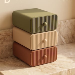 Pode incluir: Uma pilha de três gavetas de armazenamento retangulares com uma textura frontal canelada e um pequeno botão redondo em cada uma. A gaveta superior é verde-oliva, a do meio é bege e a inferior é terracota. O texto "You imagine it We print it" é visível.