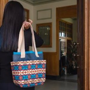 Puede incluir: Un bolso tote con un patrón geométrico vibrante en tonos azul, rojo, naranja y blanco en la parte inferior, con una banda superior azul oscuro sólida y dos asas de lona beige resistentes unidas en la parte superior para llevar al hombro.