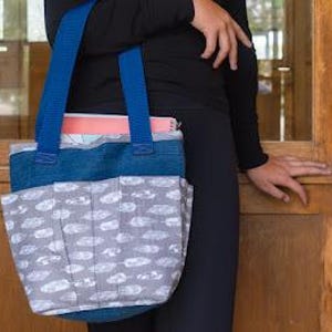 Puede incluir: Un bolso tote con una sección superior azul oscuro y una sección inferior gris con un patrón de plumas blancas. El bolso tiene dos correas azules para el hombro y un cuaderno rosa visible en el compartimento principal, ofreciendo almacenamiento.