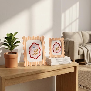 Könnte beinhalten: Zwei Bilderrahmen mit gewelltem Rand und einem vertikalen Streifenmuster in Hellorange und Weiß. Jeder Rahmen enthält eine Illustration einer weißen Blume mit blauem Zentrum auf rosa Hintergrund mit rotem Rand. Die Rahmen stehen auf Büchern.
