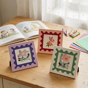 May include: Three square ceramic picture frames with floral designs. The frames are in lavender, pink, and white with scalloped edges. Each frame has a black easel back. A children's book and crayons are in the background.
