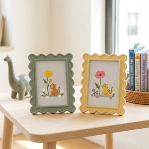May include: Two rectangular picture frames with wavy, scalloped edges. The left frame is sage green and contains an illustration of an orange cat with a yellow flower. The right frame is pale yellow and features a yellow cat with a pink flower.