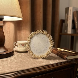 May include: A round picture frame with an ornate gold-colored metal border featuring intricate leaf and vine detailing. The frame holds a white insert decorated with a light gray shield emblem surrounded by delicate botanical line art patterns.