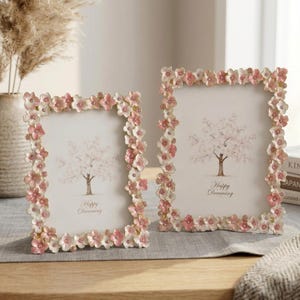 Pode incluir: Dois porta-retratos retangulares com uma borda de flores esculpidas em rosa e branco com pequenos cristais incrustados. Cada moldura contém uma inserção branca impressa com uma ilustração de árvore marrom e o texto Happy Dreaming.