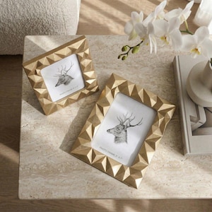 Könnte beinhalten: Zwei goldfarbene Fotorahmen mit geometrischen Mustern. Jeder Rahmen enthält ein Schwarz-Weiß-Bild eines Hirsches. Die Rahmen sind mit "PHOTO FRAME 5" x 7"" und "PHOTO FRAME 4" x 6"" beschriftet. Die Rahmen befinden sich auf einer hellen Oberfläche.