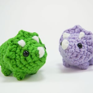 May include: Two crocheted dinosaur toys, one green and one purple, with white horns and black eyes.