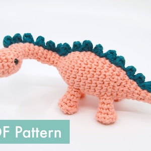 May include: A handmade crochet dinosaur in a soft peach color with teal accents. The dinosaur has a long neck, small legs, and a row of teal spikes down its back. The text "PDF Pattern" is at the bottom.