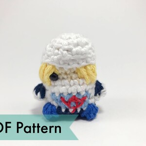 May include: A small, crocheted doll with a white hat, yellow hair, and a blue and red outfit. The doll has a black eye and is handmade. The image includes the text "PDF Pattern" on a teal banner.