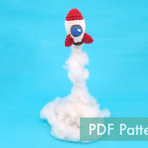 May include: A crocheted rocket toy with red, white, and blue details, launching into a white fluffy cloud against a blue background. The text "PDF Pattern" is displayed below the rocket.