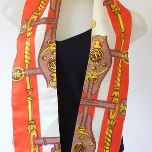 May include: A silk scarf with a horse-themed design. The scarf features a repeating pattern of brown horse tack, gold accents, and orange and white stripes. The scarf has a fringed edge.