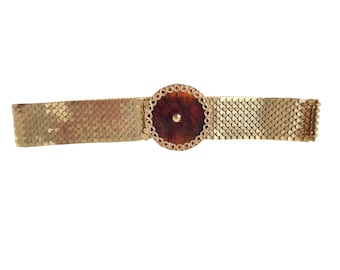 May include: A gold chain belt with a round, brown, tortoise shell buckle. The buckle has a gold rim and a small gold dot in the center.