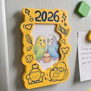 Può includere: Magnete giallo per frigorifero con l'immagine di due parrocchetti e un cupcake. Il magnete riporta l'anno 2026 in blu ed è decorato con scarabocchi. La parola "hello" è scritta sul magnete.