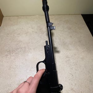 May include: Black toy gun with an extended barrel and orange tip. The gun is held in a hand, and the barrel is black with a ribbed texture. The grip and body of the gun are smooth and black.