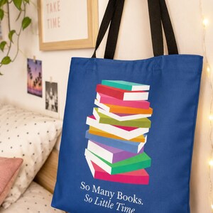 Op de afbeelding: Een koningsblauwe tote bag met zwarte schouderbanden, voorzien van een afbeelding van een kleurrijke stapel boeken en de witte tekst "So Many Books. So Little Time." op de voorzijde. De tas is gemaakt van stof en heeft een rechthoekige vorm.