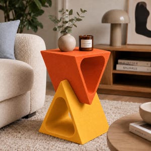 Puede incluir: Una mesa auxiliar geométrica con dos módulos triangulares apilados, la sección superior en naranja vibrante y la inferior en amarillo brillante. Un jarrón redondo beige con follaje verde y una vela marrón descansan sobre la superficie plana.