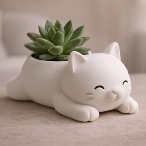 Könnte beinhalten: Ein weißer, katzenförmiger Keramik-Blumentopf mit einer grünen Sukkulentenpflanze. Die Katze liegt mit einem lächelnden Gesicht, Schnurrhaaren und Pfoten. Der Topf ist ein dekorativer Artikel für die Inneneinrichtung.