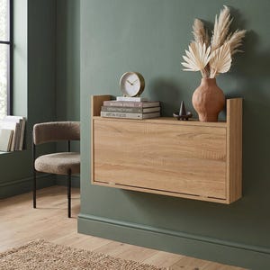 May include: A light oak wood wall-mounted drop-leaf desk featuring a rectangular surface and side panels. On top sits a round gold-rimmed clock, a stack of three books titled Modern Spaces, Geometric Forms, and Art in Focus, and a terracotta vase.