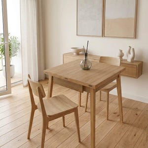 May include: A light oak dining set comprising a square table and two matching chairs. The table has a drawer and is set on a light wood floor. The room is decorated with neutral-toned artwork and decor.