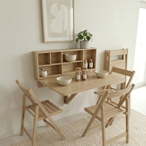 May include: A light wood-toned, wall-mounted folding table with two matching folding chairs. The table is paired with a wall-mounted shelf unit with bowls and spice jars. A framed abstract art piece and a potted plant complete the scene.