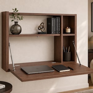 May include: A wall-mounted dark wood floating desk with a fold-down work surface supported by two metal gas struts. The unit features an upper horizontal shelf and a vertical side compartment containing books, a vase, a candle, and a pen holder.