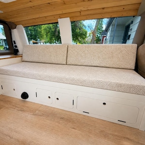 Könnte beinhalten: Innenansicht eines Vans mit einer eingebauten weißen Sitzbank und Staufächern. Die Sitzkissen sind beige mit einem kreisförmigen Muster. Der Van hat eine Holzdecke und einen hellen Holzboden. Ein Fenster zeigt die Aussicht auf eine Nachbarschaft.
