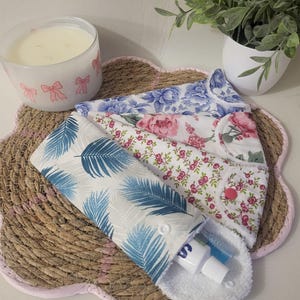 May include: A white and blue palm leaf print travel pouch with a toothbrush and toothpaste, alongside floral print fabric pads. A candle with pink bow accents and a small plant are also visible.