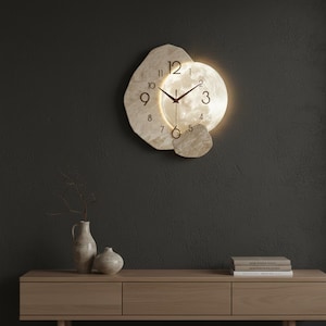 Pode incluir: Um relógio de parede moderno com uma forma orgânica única. O mostrador do relógio é bege claro com ponteiros e algarismos pretos. Uma luz circular brilhante ilumina o centro. O relógio está montado numa parede cinza escuro.