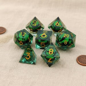 May include: A set of seven polyhedral dice in various shapes. The dice are transparent green with gold numbering and a green eye design inside. The dice are arranged on a neutral surface.