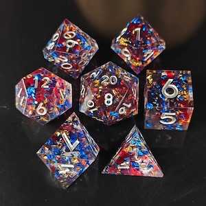 Puede incluir: Juego de siete dados poliédricos para juegos de rol. Los dados transparentes contienen inclusiones rojas, azules y doradas, con números plateados. Cada dado tiene un número diferente de caras.