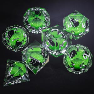 May include: A set of seven polyhedral dice with a green glitter interior and black cat illustrations. The dice have silver numbering and are made of clear resin. The dice are designed for tabletop role-playing games.