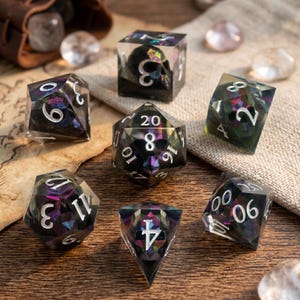 May include: A collection of seven polyhedral dice, ideal for role-playing games. The dice are black with shimmering, multi-colored inclusions and white numerals. One die has a skull. The dice are on a wooden surface.