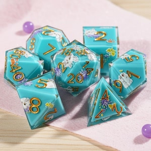 Könnte beinhalten: Ein Satz von sieben polyedrischen Würfeln in verschiedenen Formen. Die Würfel haben eine durchscheinende türkisfarbene Farbe mit goldenen Zahlen und Katzenillustrationen. Florale Akzente in Lila und Gelb sind ebenfalls vorhanden. Eine lila Perle befindet sich im Hintergrund.