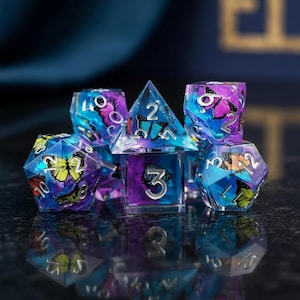 May include: A set of iridescent dice with butterfly designs. The dice are clear with blue, purple, and yellow butterflies inside. The numbers are silver. The dice are arranged on a dark, reflective surface.